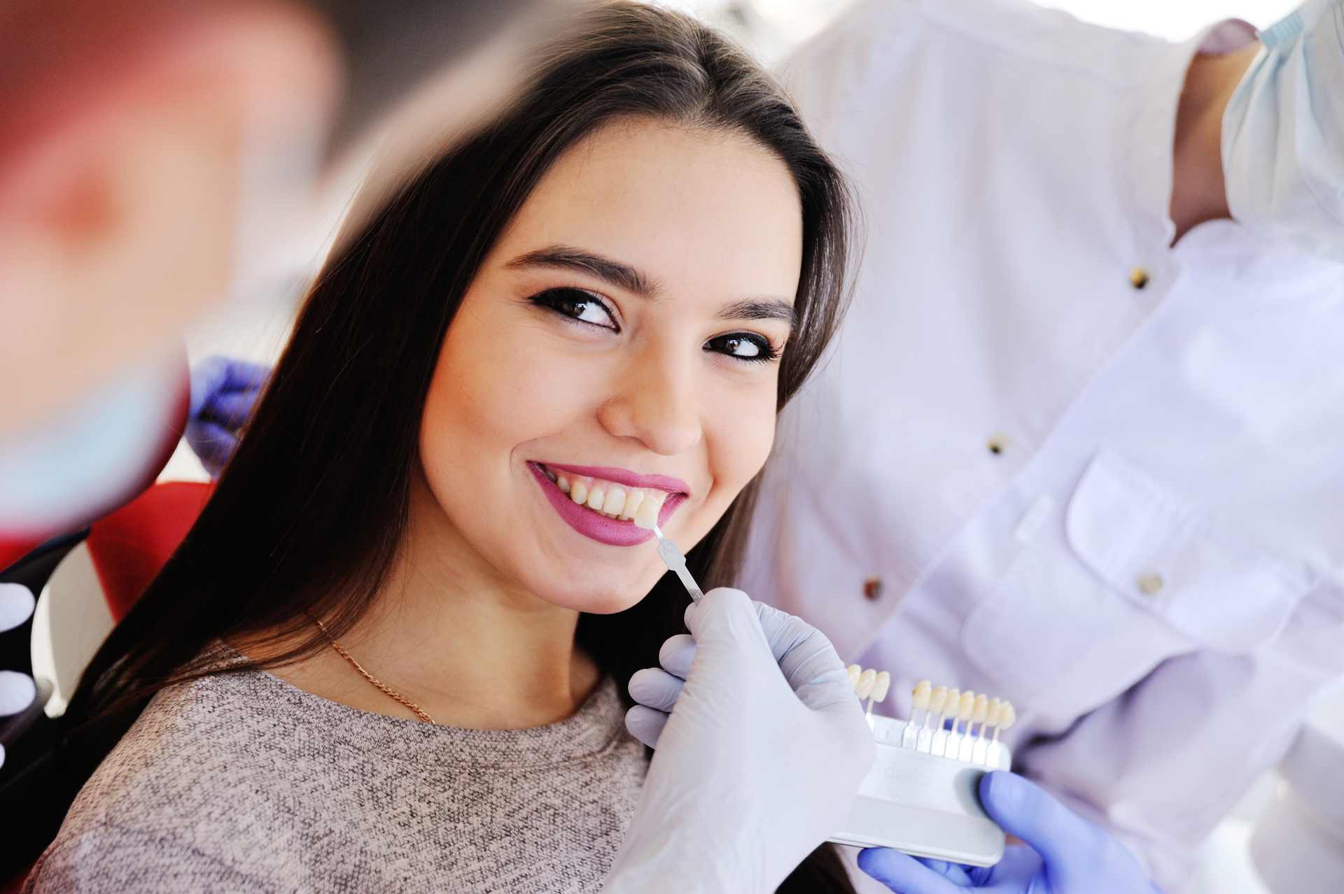 01 Dentist helping a smiling female patient choose from the porcelain veneer color chart_Lumineers vs. porcelain veneers